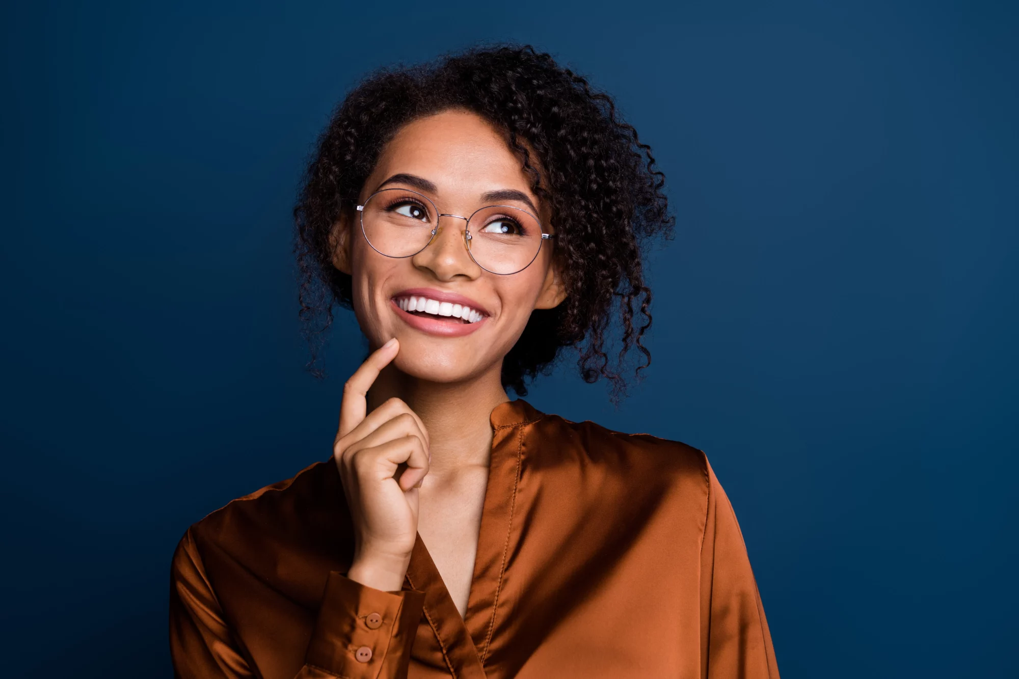 Smiling woman with curly hair and glasses, hand on her chin, against a blue backdrop